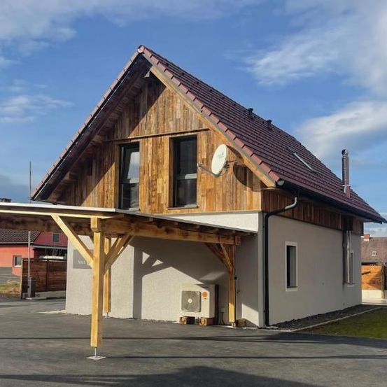 Ein modernes Holzhaus mit überdachtem Parkplatz und einer Satellitenschüssel auf dem Dach. Das Haus ist weiß mit braunen Verkleidungen, und es hat ein rotes Dach.
