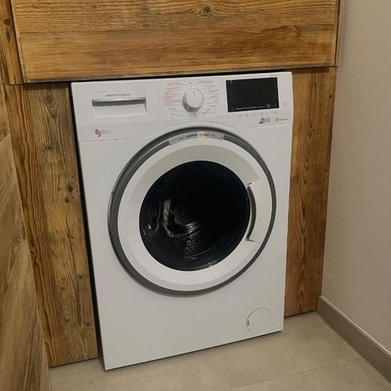 Eine weiße Waschmaschine mit einer grauen Tür steht auf dem Boden in einem Raum. Die Waschmaschine hat ein digitales Display. Der Raum hat Holzwände und gefliesten Boden.