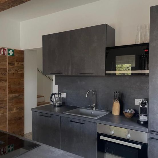 Moderne Küche mit Holzschränken und einer Spüle. Die Arbeitsplatte ist grau und der Ofen und die Mikrowelle sind an der Wand. Eine Kaffeemaschine befindet sich rechts.