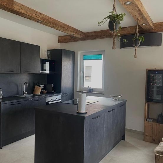 Eine moderne Küche mit einer zentralen Insel, schwarzen Schränken und einer Holzdecke. Die Küche hat ein Waschbecken, einen Herd und eine Mikrowelle. Es gibt ein Fenster mit Blick nach draußen.