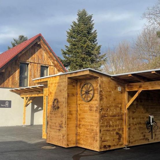 Ein Holzgebäude mit rotem Dach und einem Rad an der Wand steht neben einem größeren Holzhaus mit grauer Wand.