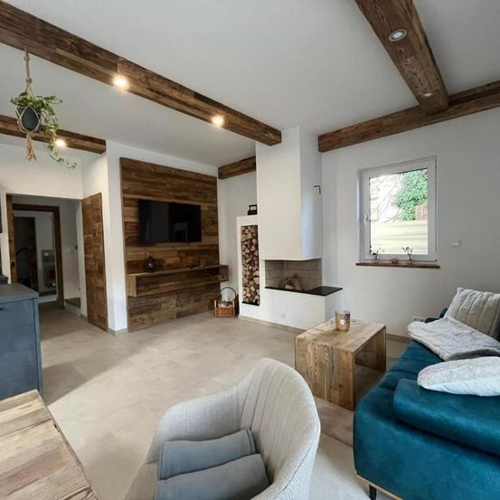 Ein modernes Wohnzimmer mit Holzbalken, einem Kamin und einem Flachbildfernseher. Der Raum hat einen Holzboden, ein blaues Sofa mit Kissen, einen Holztisch und einen Stuhl.