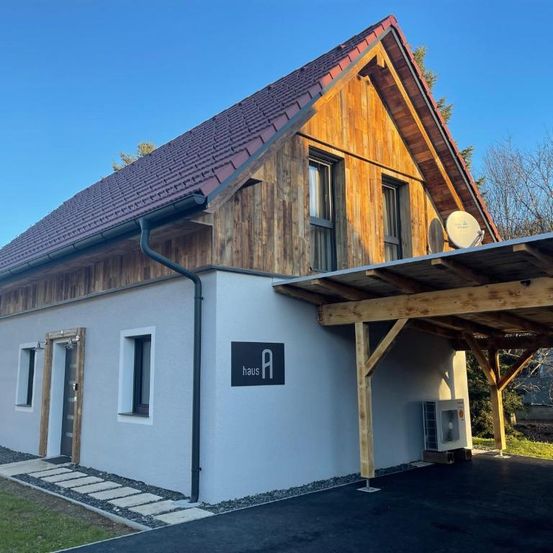 Ein holzverkleidetes und weiß gestrichenes Haus mit rotem Dach und einer Satellitenschüssel. Es hat zwei Fenster links und eine hölzerne Pergola rechts.