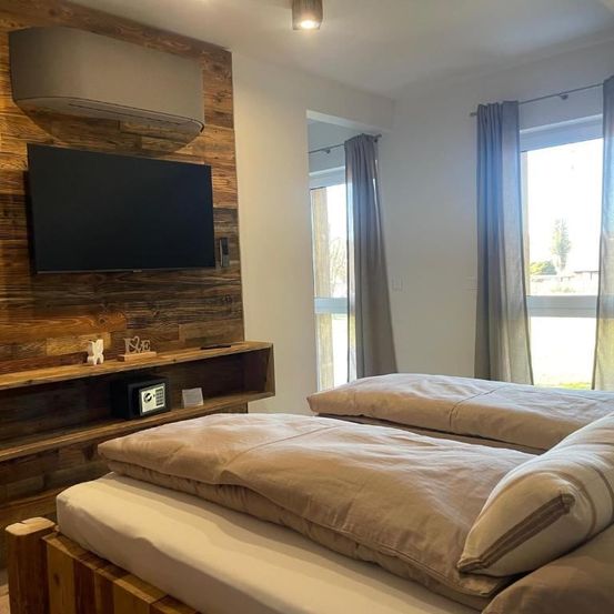 Ein Schlafzimmer mit zwei Betten und einer Holzwand. Ein Flachbildfernseher ist an der Wand montiert und ein Regal hält einen digitalen Safe. Das Zimmer hat Fenster mit Vorhängen.