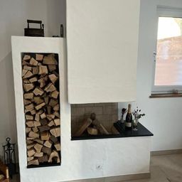 Ein leerer Kamin mit einem Stapel Holzscheite links und einem Regal mit einer Flasche Champagner, Blumen und einem Weinglas rechts.