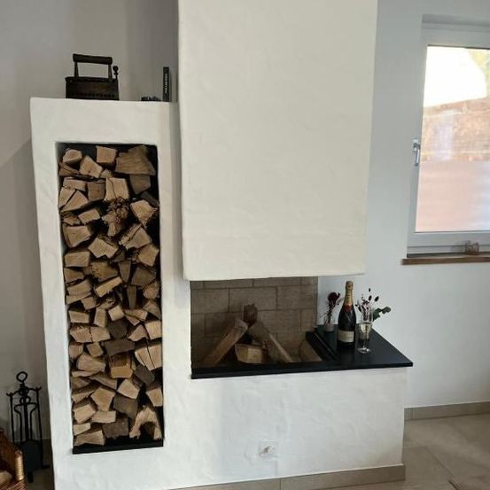 Ein leerer Kamin mit einem Stapel Holzscheite links und einem Regal mit einer Flasche Champagner, Blumen und einem Weinglas rechts.
