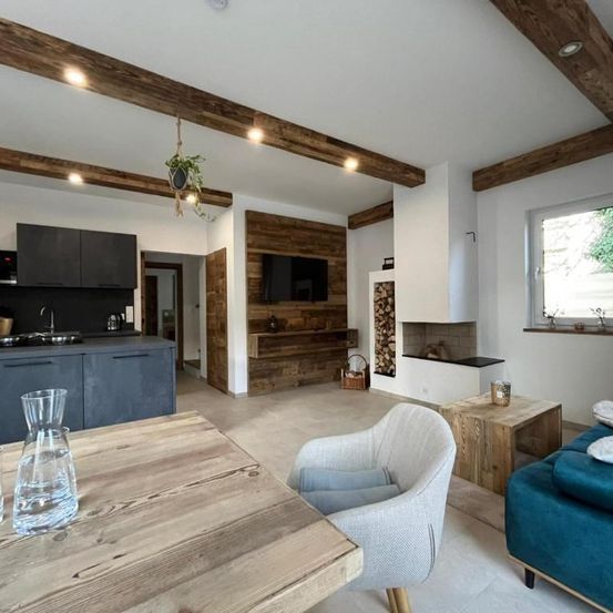 Ein modernes Wohnzimmer mit Holzbalken, einem Kamin und einem blauen Sofa. Im Vordergrund steht ein Holztisch mit einem Glaskrug.