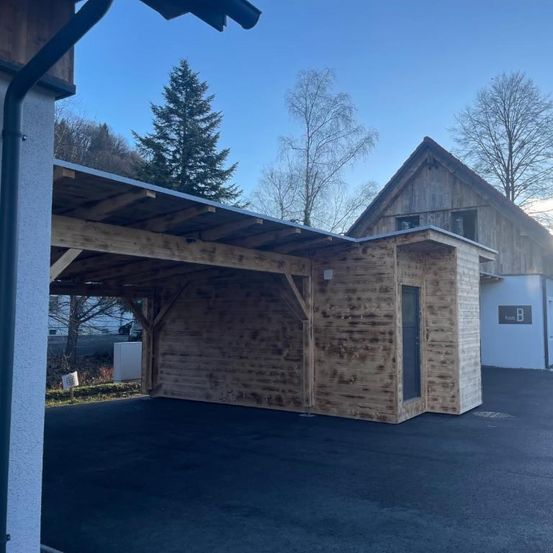 Ein hölzerner Carport mit einem geneigten Dach steht vor einem Holzhaus. Bäume umgeben das Gebiet.