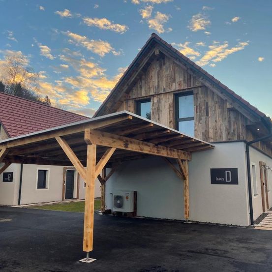 Ein Holzhaus mit einem Carport unter blauem Himmel und Wolken, mit einem roten Dach und einer weißen Außenfassade. Das Haus hat Glasfenster und ein Schild, das 'Haus D' steht. Es gibt einen schwarzen Carport, eine Klimaanlage an der Wand und einen Weg.