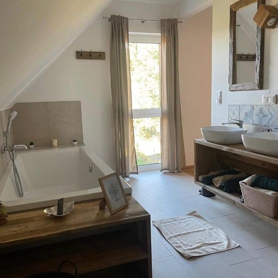 Ein Badezimmer mit Badewanne, Dusche, zwei Waschbecken und einem Spiegel. Der Raum verfügt über Holzregale, weiße Fliesen und beige Vorhänge.