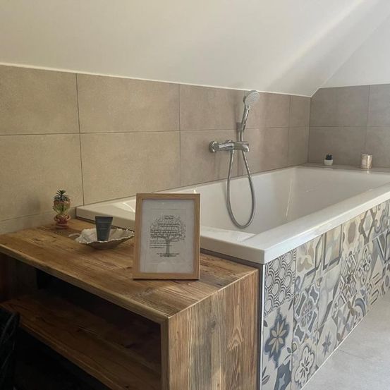 Ein hölzerner Regal neben einer Badewanne mit einer Duschkopf. Ein kleiner Topf und ein Bilderrahmen sind auf dem Regal. Die Badewanne hat geflieste Wände.