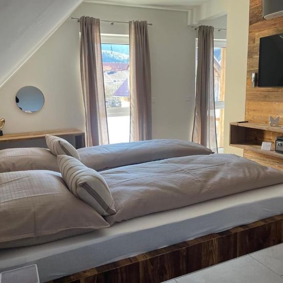 Ein Schlafzimmer mit zwei Betten, jedes mit zwei Kissen und Decken, ist zu sehen. Ein Spiegel und ein Fernseher sind an den Wänden angebracht, und Vorhänge bedecken die Fenster.