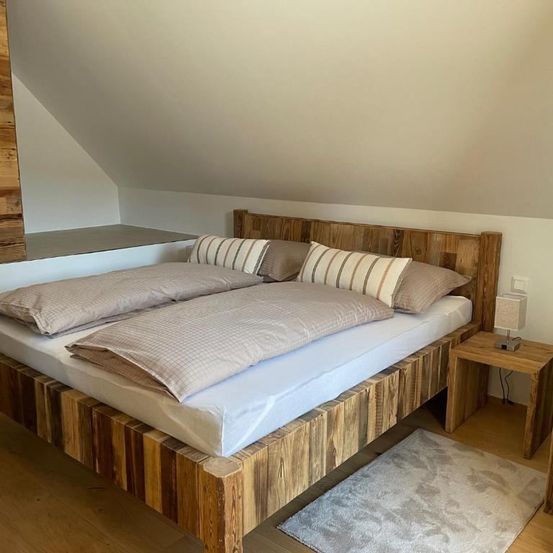 Ein gemütliches Schlafzimmer mit Holzbett, Kissen und Nachttisch mit Lampe an einer attikaartigen Holzwand.