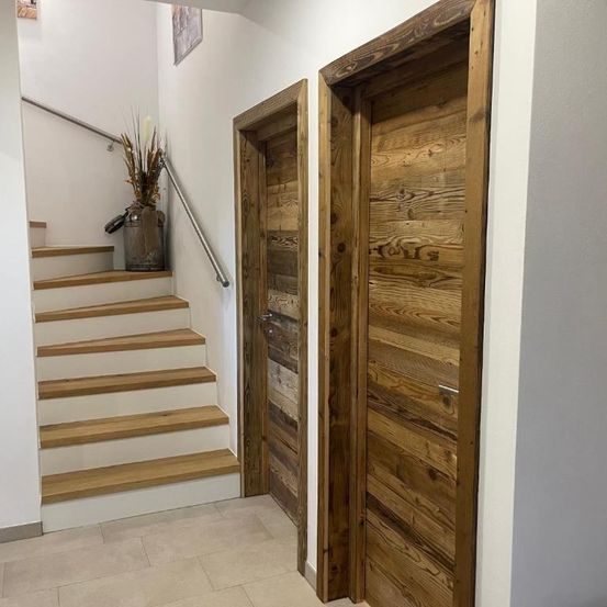 Innenansicht eines Hauses mit Holztreppen und zwei Holztüren auf beiden Seiten. Ein Topf mit einer Pflanze steht auf der Treppe.