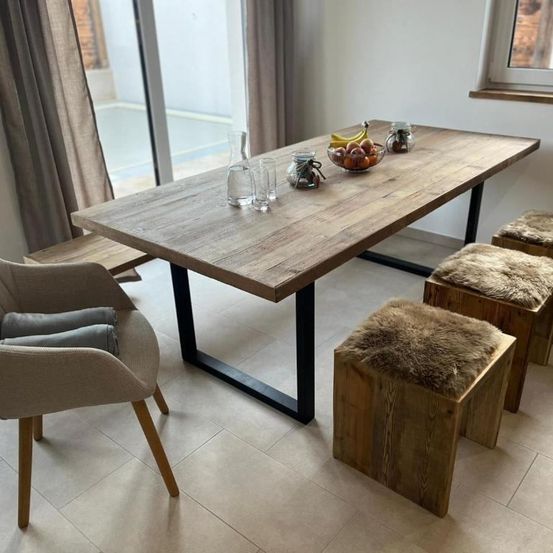 Ein hölzerner Esstisch mit einer Schüssel Obst darauf, umgeben von Holzhockern mit Fellauflagen, einer Bank und einer Glastür mit Blick auf eine Ziegelmauer.
