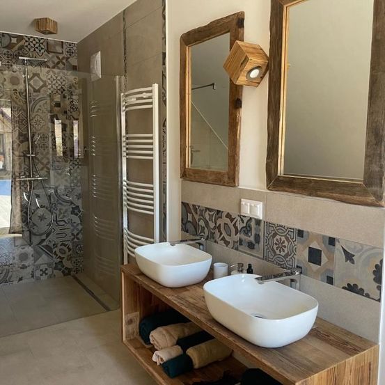 Ein Badezimmer mit gefliesten Wänden, einer Dusche und einem Holztresen mit zwei Waschbecken. Es gibt zwei Spiegel und einen Handtuchhalter.