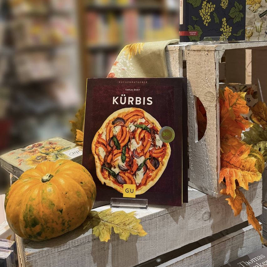 Ein Kochbuch mit dem Titel 'Kurbis' zeigt eine Pizza auf dem Cover, auf einem Holzregal mit Herbstblättern und einer Kürbis.