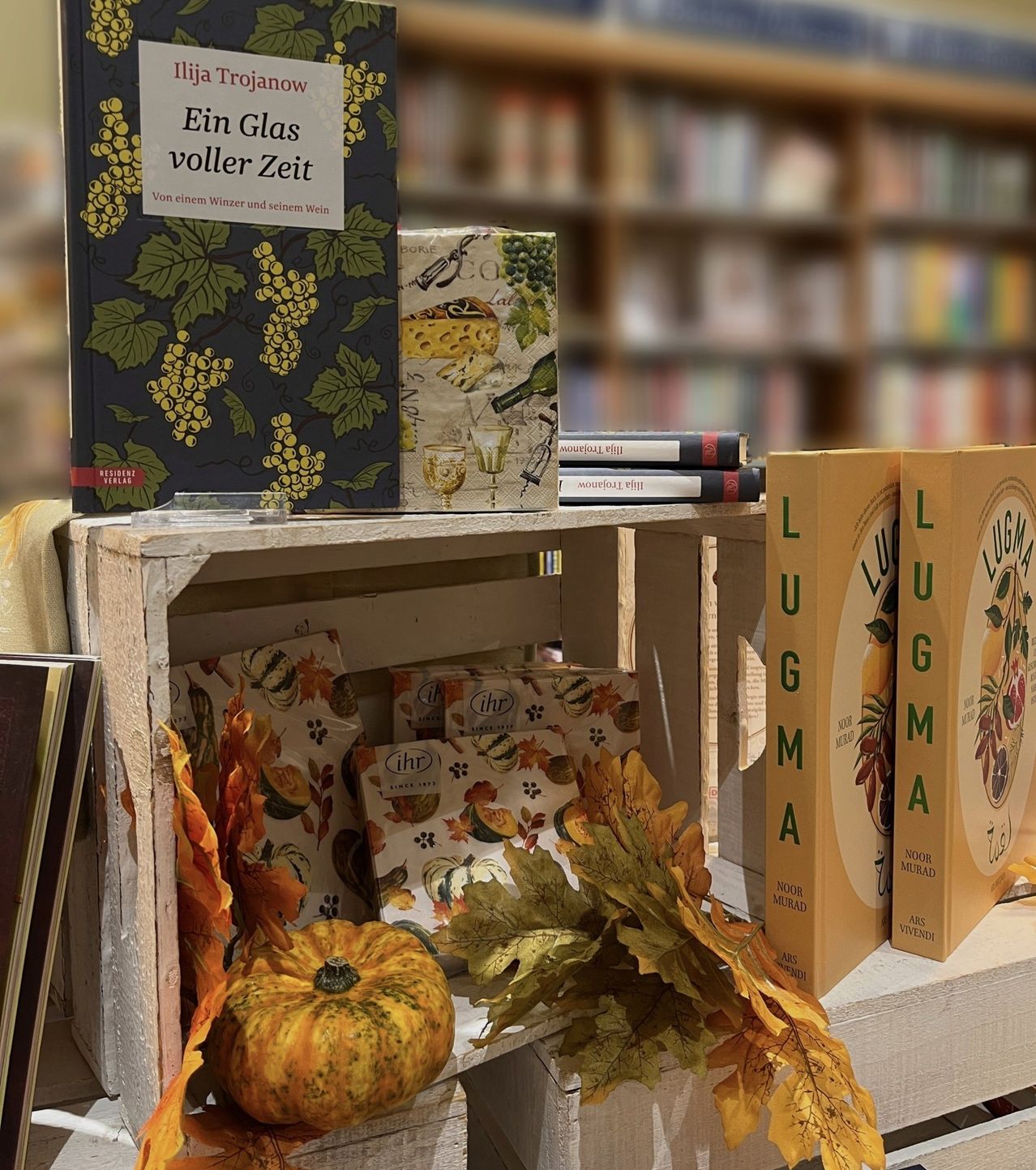 Ein Holzkisten mit Büchern, Kürbissen und Herbstblättern. Die Bücher haben Titel wie Ein Glas voller Zeit und LUGMA. Der Hintergrund zeigt eine Bibliothek mit Bücherregalen.