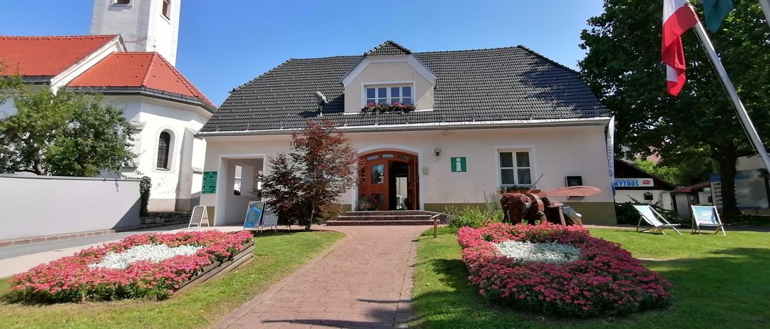 Ein weißes Gebäude mit grauem Dach und einem Glockenturm steht neben einem großen Baum. Es hat Blumen und eine Holztür. Ein Weg mit Blumen führt zum Eingang.
