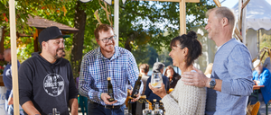 Eine Gruppe von Menschen, die lächeln und sich unterhalten, steht an einem Tisch mit Flaschen und Gläsern. Eine Frau hält ein Glas Wein und ein Mann neben ihr eine Flasche. Dahinter stehen weitere Personen und Bäume.