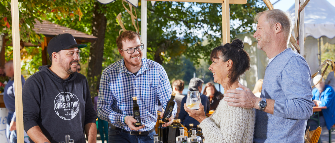 Eine Gruppe von Menschen, die lächeln und sich unterhalten, steht an einem Tisch mit Flaschen und Gläsern. Eine Frau hält ein Glas Wein und ein Mann neben ihr eine Flasche. Dahinter stehen weitere Personen und Bäume.