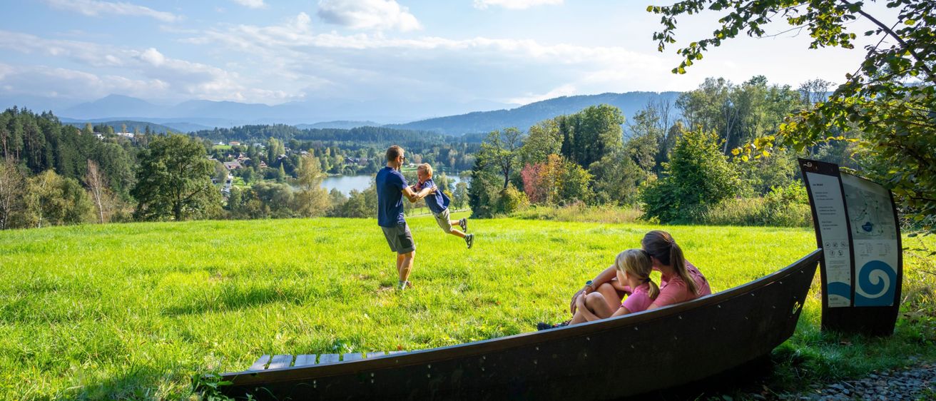 Eine Familie genießt einen sonnigen Tag auf einer Wiese mit einem See und Bergen im Hintergrund.