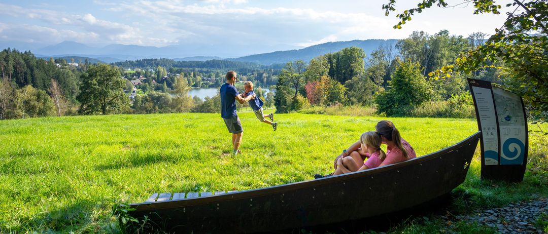Eine Familie genießt einen sonnigen Tag auf einer Wiese mit einem See und Bergen im Hintergrund.