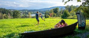Eine Familie genießt einen sonnigen Tag auf einer Wiese mit einem See und Bergen im Hintergrund.