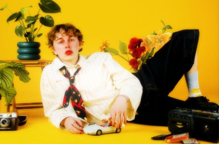 Ein junger Mann liegt auf einem gelben Hintergrund, hält ein Spielzeugauto und ist von Pflanzen und einer Kamera umgeben.