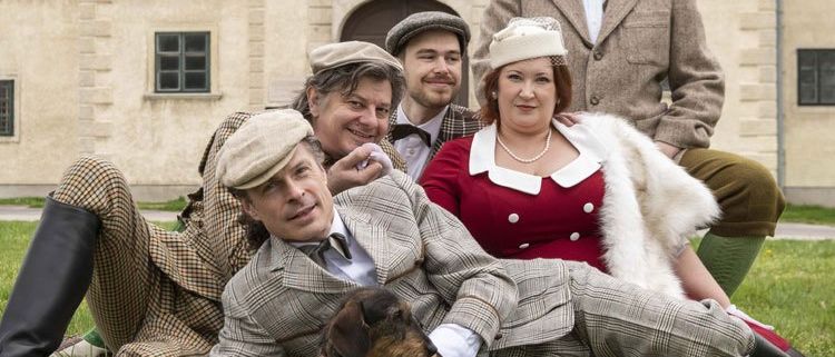 Eine Gruppe von fünf Personen in Vintage-Kleidung posiert auf einem Rasen vor einem historischen Gebäude.