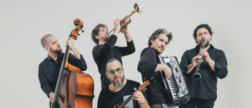 Fünf Musiker in Schwarz, posieren mit ihren Instrumenten. Einer sitzt auf einem Stuhl, während andere hinter ihm stehen. Sie alle schauen zur Kamera.