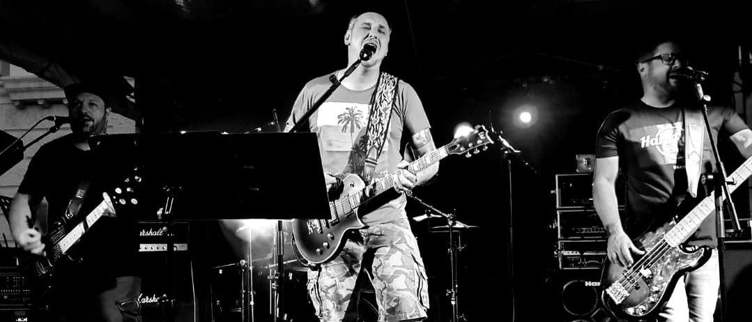 Ein Schwarz-Weiß-Foto einer Band, die auf der Bühne vor einem Mikrofon auftritt. Der Mann in der Mitte singt und spielt Gitarre. Die anderen spielen Bassgitarre und Schlagzeug.