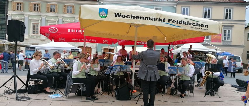 Ein Orchester spielt auf einem Stadtplatz unter einem Schirm mit der Aufschrift 'Wochenmarkt Feldkirchen'. Mehrere Musiker sitzen auf Stühlen und spielen verschiedene Instrumente, während ein Dirigent vor ihnen steht. Im Hintergrund befinden sich Gebäude mit roten Dächern und weißen Fenstern.