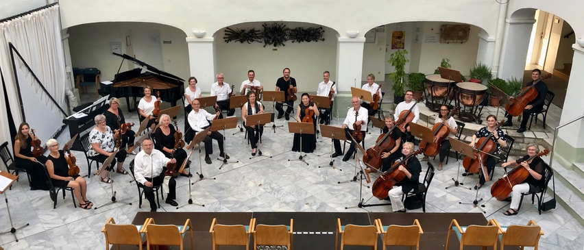 Ein klassisches Musikensemble spielt Instrumente, darunter Violinen, Celli und ein Akkordeon, in einem Raum mit gefliestem Boden und weißen Wänden. Die Musiker sitzen auf Stühlen, einige stehen.