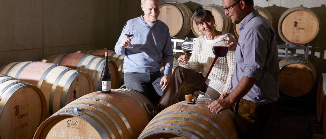 Drei Personen probieren Wein in einem Weinkeller, umgeben von Fässern. Die Frau schenkt Wein aus einer Flasche ein, während die beiden Männer Gläser halten. Die Fässer sind mit verschiedenen Texten und Zahlen beschriftet.
