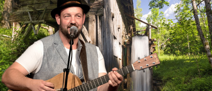 Ein Mann in Hut und Weste spielt Gitarre an einem Wasserfall unter einer alten Holzbrücke an einem sonnigen Tag.