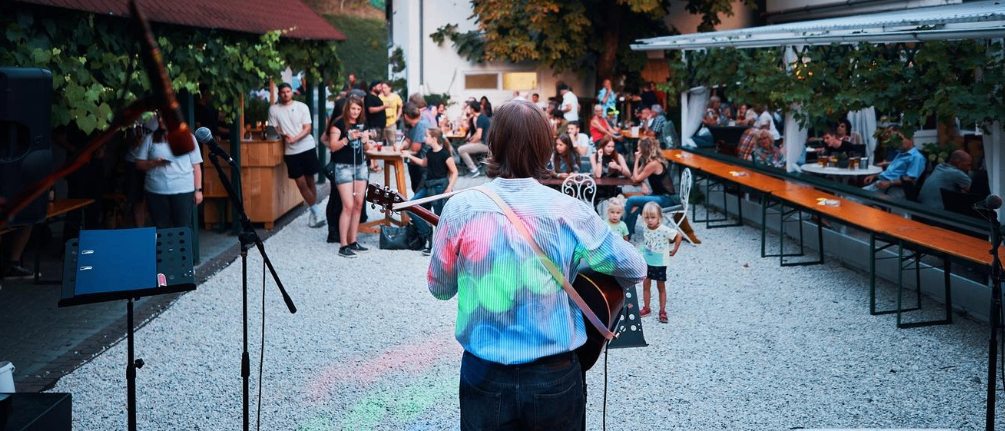 Ein Gitarrist tritt auf einer Bühne in einem lebhaften Außenbereich auf. Eine Menge versammelt sich und genießt die Musik. Die Szene umfasst Tische, Stühle und einen Baum, der Schatten spendet.