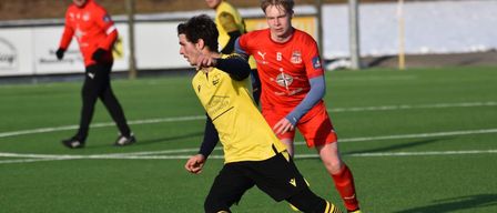 Two soccer players in yellow and red uniforms play on a field, one dribbling a soccer ball.