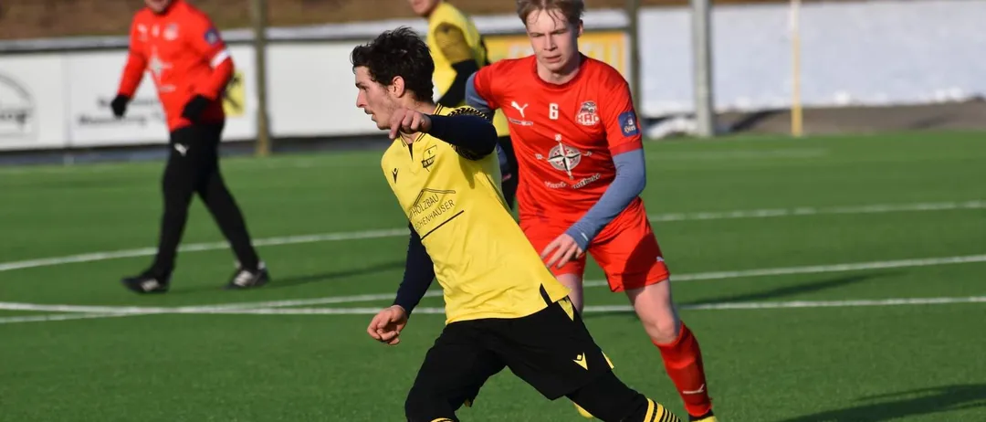 Zwei Fußballspieler in gelben und roten Trikots spielen auf einem Feld, einer dribbelt einen Fußball.