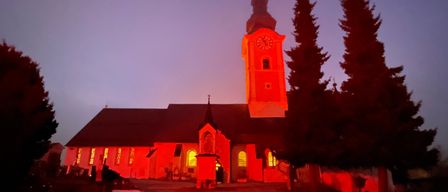 Beim Einbruch der Dunkelheit wird eine Kirche mit rotem Licht beleuchtet und zeigt einen hohen Glockenturm, einen Dachreiter und mehrere Rundbogenfenster.
