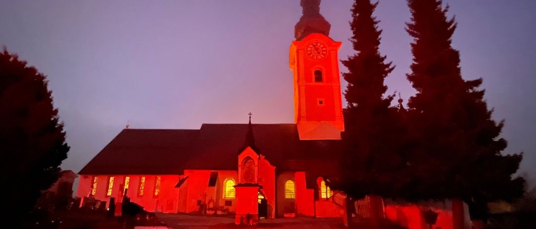 Beim Einbruch der Dunkelheit wird eine Kirche mit rotem Licht beleuchtet und zeigt einen hohen Glockenturm, einen Dachreiter und mehrere Rundbogenfenster.