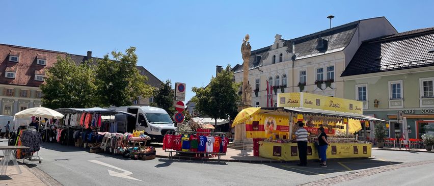 Ein Outdoor-Markt mit verschiedenen Ständen unter Sonnenschirmen und Markisen ist in der Nähe einer Statue aufgebaut. Ein weißer Van ist in der Nähe geparkt. Menschen stehen an einem Stand mit gelben Bannern. Gebäude mit vielen Fenstern sind im Hintergrund sichtbar.