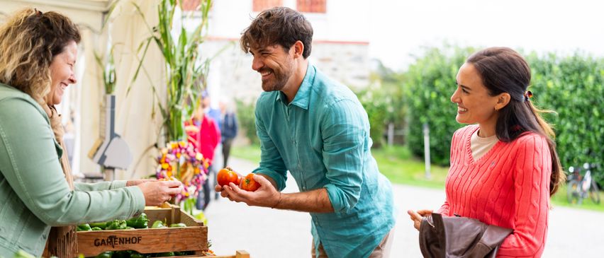 Ein Mann, der Tomaten hält, steht lächelnd an einem Marktstand mit einer Frau in einem roten Pullover und einer anderen Person, die nach Gemüse greift.