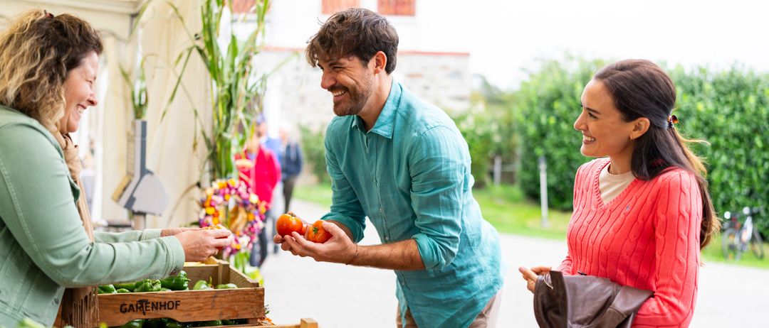 Ein Mann, der Tomaten hält, steht lächelnd an einem Marktstand mit einer Frau in einem roten Pullover und einer anderen Person, die nach Gemüse greift.