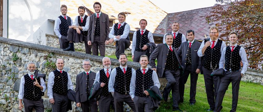 Eine Gruppe von Männern in traditioneller Kleidung, die auf einer Steinmauer stehen und sitzen. Sie tragen rote Krawatten und Westen mit weißen Punkten. Im Hintergrund befinden sich Gebäude und Grünflächen.