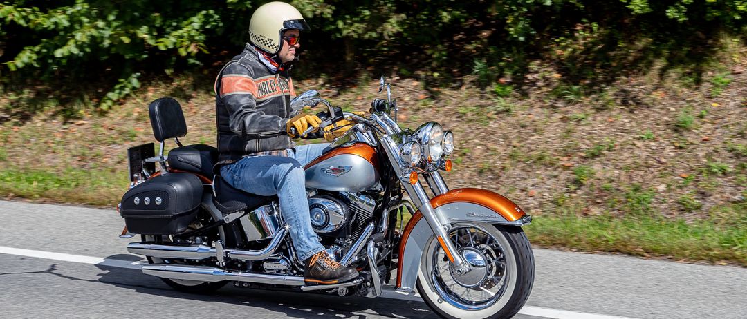 Ein Mann fährt mit einem Helm, Sonnenbrille und Handschuhen eine Harley-Davidson-Motorrad auf der Straße. Das Motorrad hat ein Chrom- und Orange-Farbschema.
