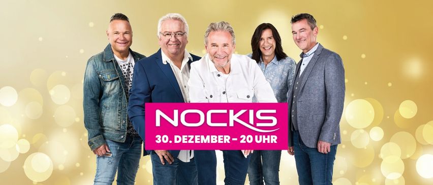 Fünf Personen stehen eng beieinander und lächeln. Ein rosa Schild mit dem Text 'NOCKIS 30. DEZEMBER - 20 UHR' steht in der Mitte. Der Hintergrund ist gelb.