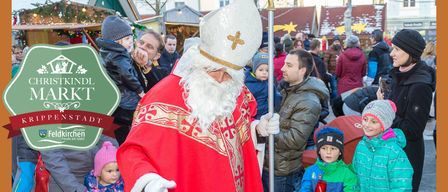 Eine Menschenmenge versammelt sich um den Weihnachtsmann auf einem Weihnachtsmarkt und hält rote Geschenktüten. Die Szene ist lebhaft mit mehreren Menschen in festlicher Winterkleidung. Gebäude im Hintergrund sind mit Weihnachtsdekorationen geschmückt.