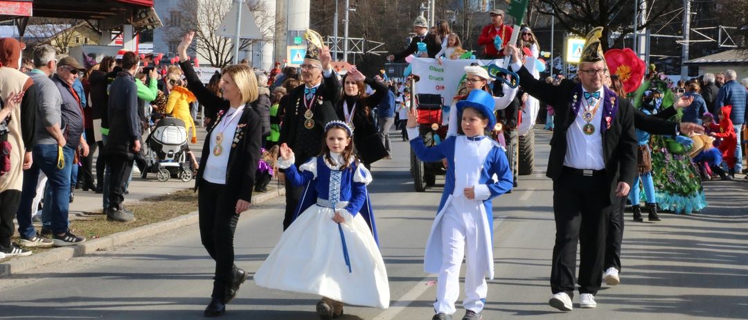 Ein Umzug mit mehreren Teilnehmern in Kostümen. Eine Frau und ein Kind in blau-weißen Outfits gehen auf der Straße. Ein erwachsener Mann trägt Hut und Brille.