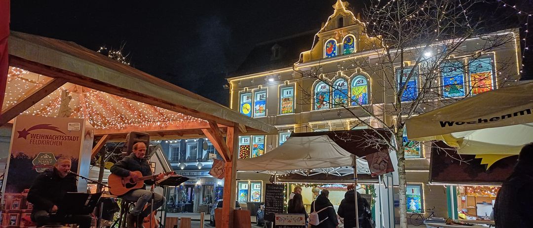 Ein Musiker spielt Gitarre auf der Bühne bei Nacht, mit einem Publikum in der Nähe. Ein Zelt ist neben einem Gebäude mit bunten Fenstern aufgestellt. Lichter und Dekorationen sind zu sehen.